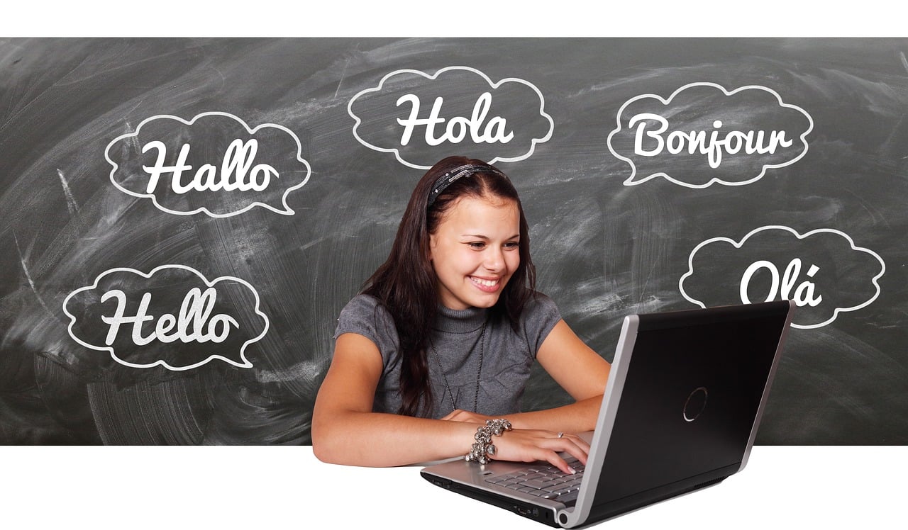 Foto con una pizarra que contiene la palabra hola en varios idiomas y una alumna con un ordenador