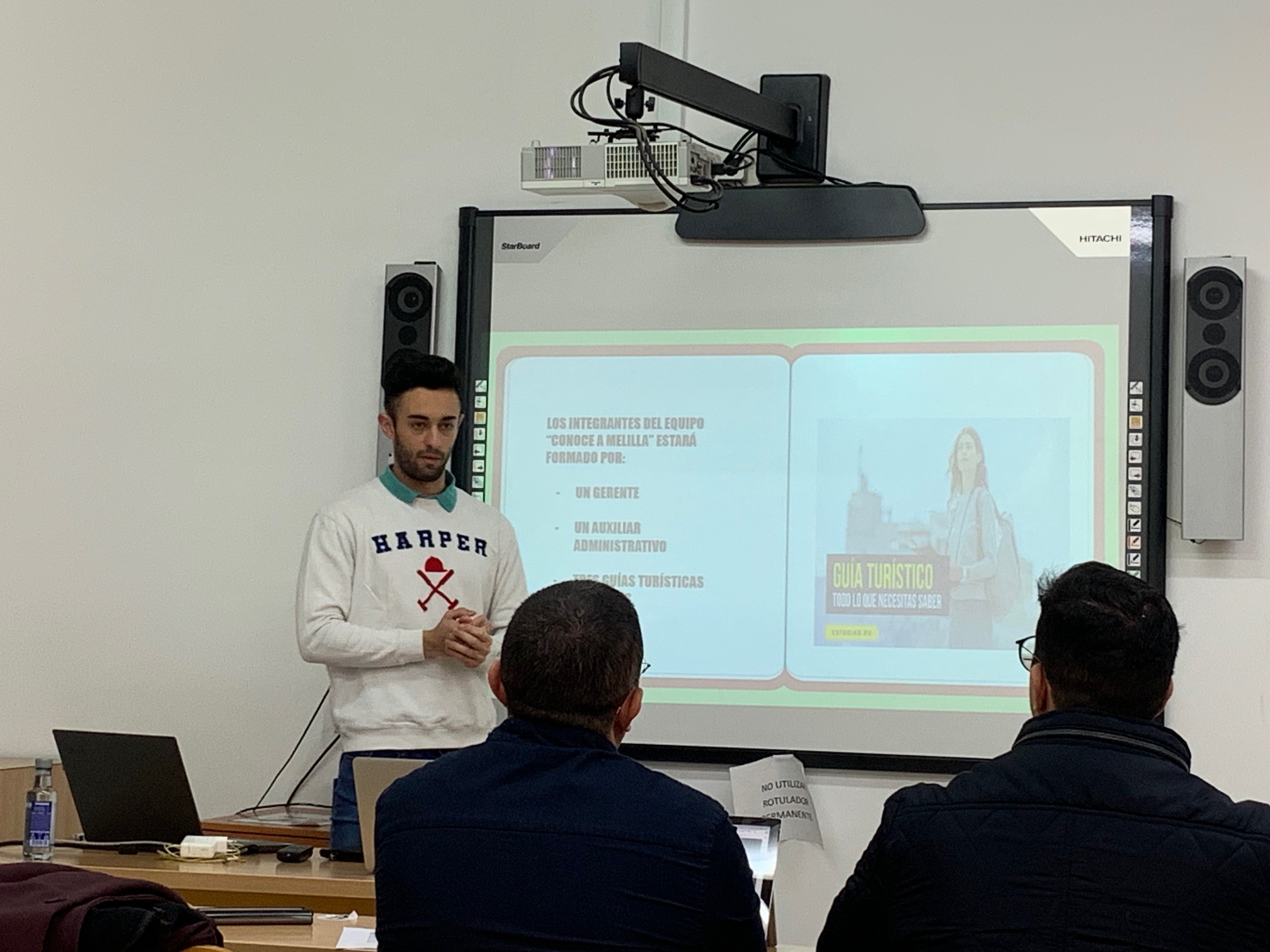 Foto del ganador explicando su idea con una presentación en una pantalla a dos examinadores que están sentados frente a él