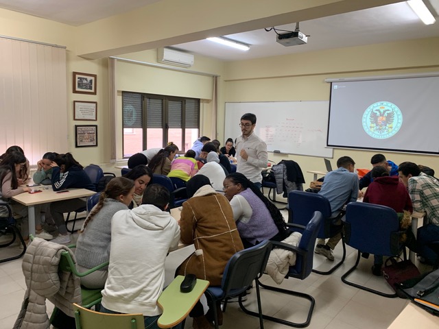 Foto de la clase con alumnos por grupos y el profesor explicando