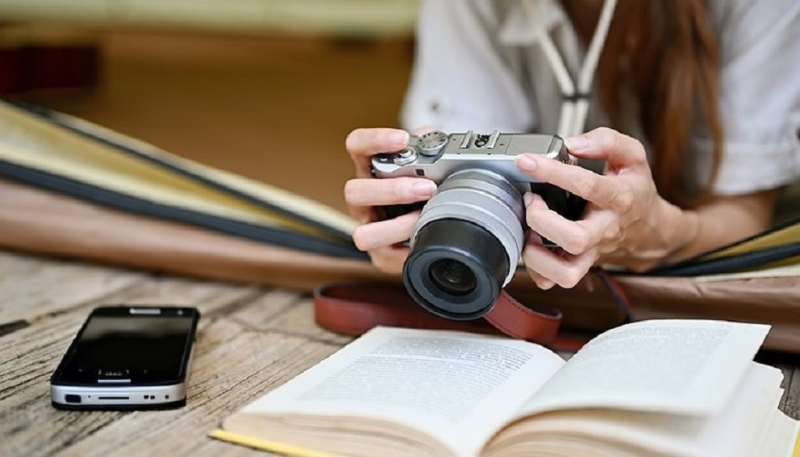 II Concurso de Fotografía “FACISO en Melilla” cámara de fotos sostenida por una mujer sobre un escritorio con un libro de texto y un móvil